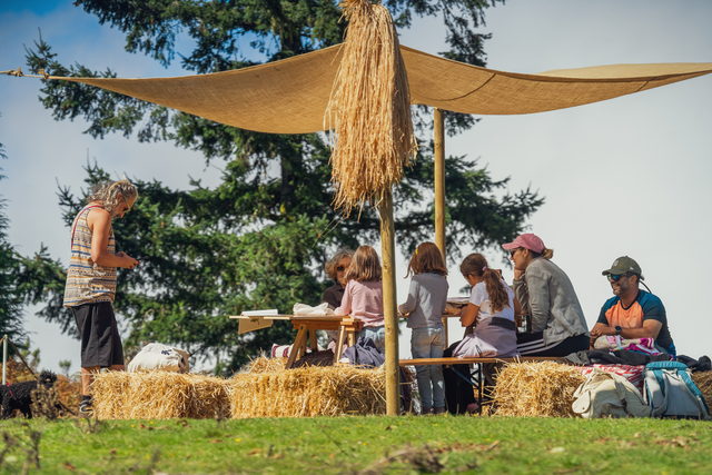 Crianças e famílias a desenhar numa mesa de madeira, sob um toldo de serapilheira, entre fardos de palha