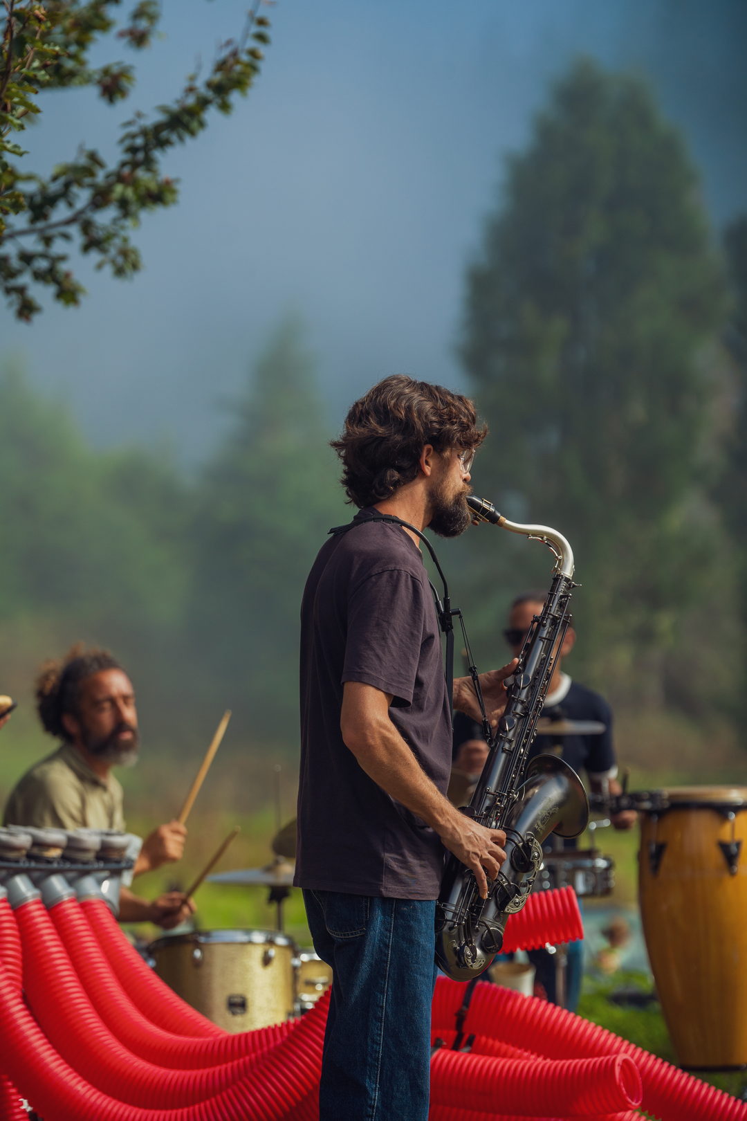 Saxofonista e percussionista a tocar ao ar livre, com o nevoeiro a subir por trás das árvores.