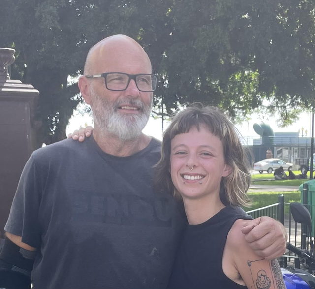 Pai e filha abraçados e a sorrir ao ar livre — ele de barba branca e óculos, ela com o braço tatuado — com árvores atrás.