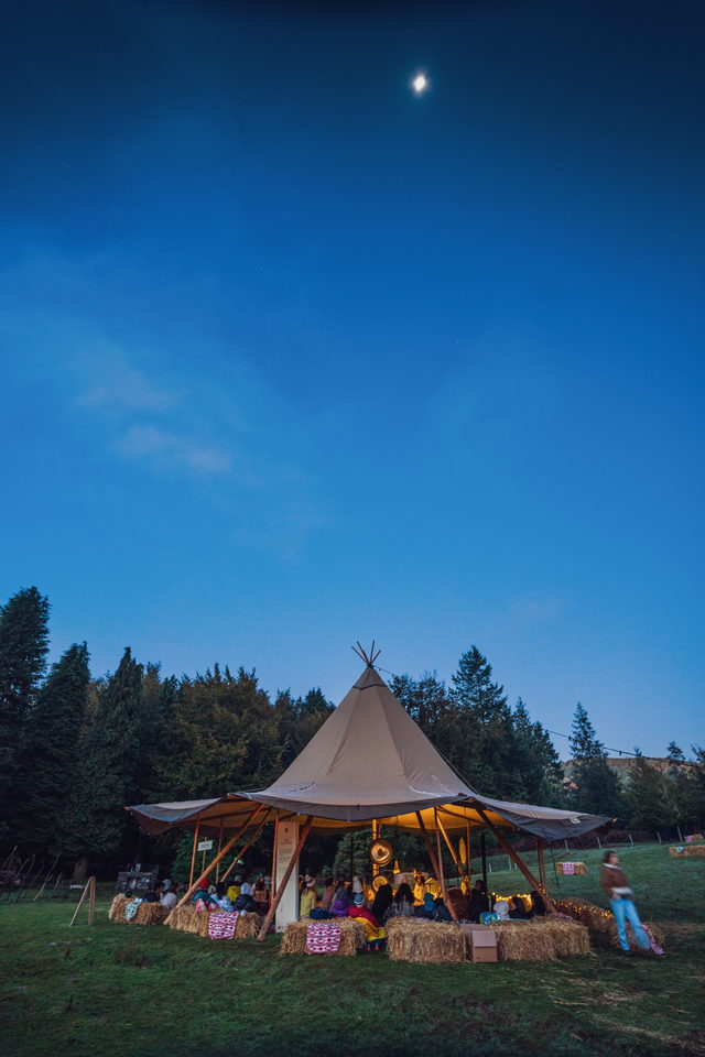 Tenda tipi iluminada por dentro ao anoitecer, com pessoas sentadas em fardos de palha e a lua no céu azul